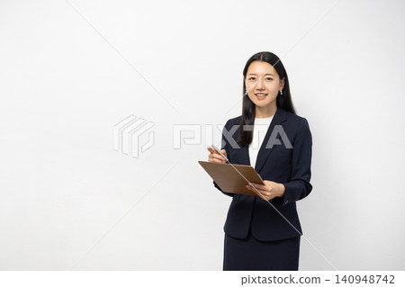 一位年輕的日本女商人對著鏡頭微笑，同時在白牆前的活頁夾上做筆記。 140948742