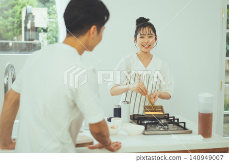 A man watches over a woman cooking tamagoyaki (rolled omelet) for a bento box, and the scene is set in a kitchen. A man watches over a woman cooking tamagoyaki (rolled omelet) for a bento box, and the scene is set in a kitchen. 140949007