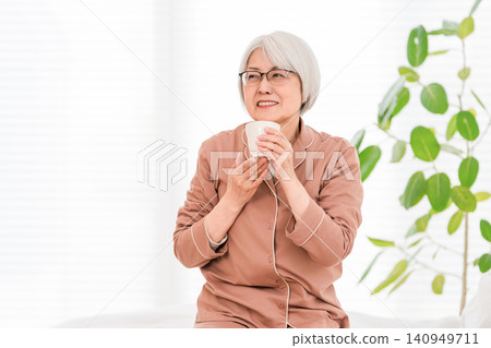 一位頭髮花白的老婦人,穿著睡衣,起床後在臥室補充水分。 一位頭髮花白的老婦人,穿著睡衣,起床後在臥室補充水分。 140949711