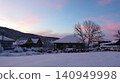 A snow-covered village in the morning or evening twilight. A snow-white field stretches in the foreground, houses with snow-covered roofs are visible beyond, and the sky is painted in soft pink hues. 140949998