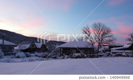 A snow-covered village in the morning or evening twilight. A snow-white field stretches in the foreground, houses with snow-covered roofs are visible beyond, and the sky is painted in soft pink hues. A snow-covered village in the morning or evening twilight. A snow-white field stretches in the foreground, houses with snow-covered roofs are visible beyond, and the sky is painted in soft pink hues. 140949998