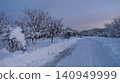 A cleared country road in the winter twilight. High snowdrifts line the roadsides, and the trees and bushes are thickly covered with a fluffy crust. The sky on the horizon is painted in soft pink tone 140949999