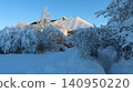A country house with a huge snow cap on its roof on a winter day. In front of the building is a snow-covered garden with bushes under a heavy crust, and to the right is a cleared narrow path. 140950220