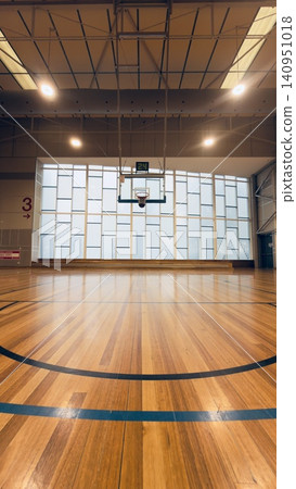 A vertical low-angle perspective of an empty indoor basketball court with shiny hardwood flooring, looking towards the hoop under bright ceiling lights and a large geometric window background. A vertical low-angle perspective of an empty indoor basketball court with shiny hardwood flooring, looking towards the hoop under bright ceiling lights and a large geometric window background. 140951018