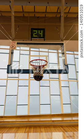 A vertical close-up shot of a unique blue and yellow basketball falling through the net of a basketball hoop, capturing the scoring moment in an indoor gym with a geometric window background. A vertical close-up shot of a unique blue and yellow basketball falling through the net of a basketball hoop, capturing the scoring moment in an indoor gym with a geometric window background. 140951019