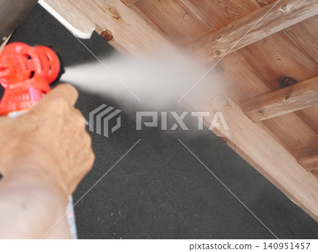 An elderly person's hand sprays a chemical onto the roofing in the attic with a red spray bottle. An elderly person's hand sprays a chemical onto the roofing in the attic with a red spray bottle. 140951457