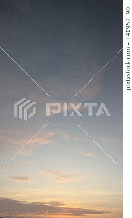 A beautiful vertical dawn landscape view captures soft pink tinted clouds drifting across a peaceful pale blue pastel sky during a calm sunrise. The gentle gradient color transition creates a serene 140952190