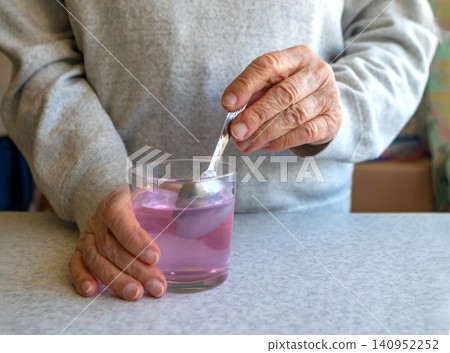 Elderly person stirring pink liquid medicine in a glass with a spoon at kitchen table 140952252