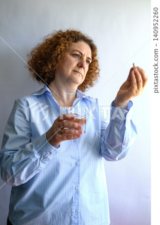 Middle-aged woman considering medication while holding a pill and glass of water at home 140952260