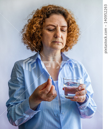 Middle-aged woman taking a pill with a glass of water, thoughtful health routine at home Middle-aged woman taking a pill with a glass of water, thoughtful health routine at home 140952263