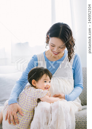 一個小女孩依偎在她懷孕的母親身邊,兩人在客廳玩耍。 一個小女孩依偎在她懷孕的母親身邊,兩人在客廳玩耍。 140952504