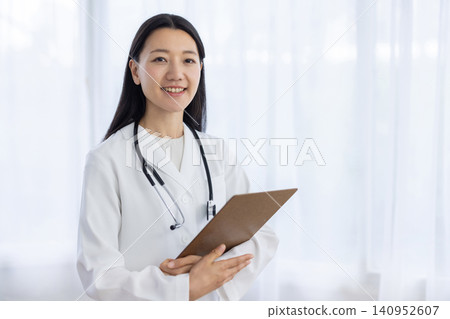 A young Japanese female doctor smiles at the camera, holding a binder, by the window. A young Japanese female doctor smiles at the camera, holding a binder, by the window. 140952607