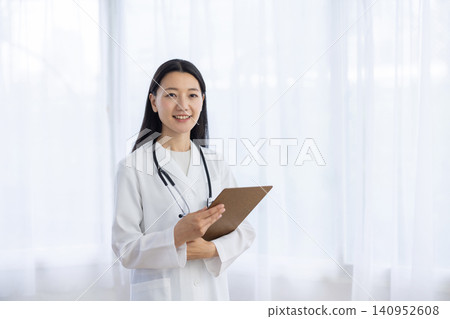 A young Japanese female doctor smiles at the camera, holding a binder, by the window. A young Japanese female doctor smiles at the camera, holding a binder, by the window. 140952608