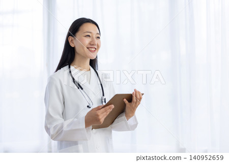 一位年輕的日本女醫生面帶微笑地站在窗邊，手裡拿著一個文件夾。 140952659