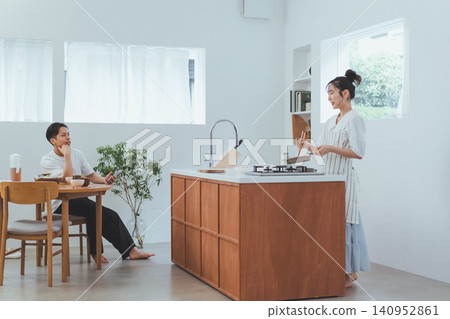 A bright living room/dining room/kitchen (LDK) scene where a man talks to his girlfriend while she is cooking from the dining room. 140952861