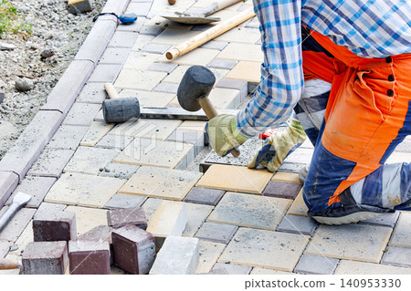 Worker lays down paving stones at a construction site in the afternoon, using a rubber mallet to ensure a strong fit 140953330