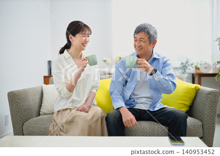 A smiling couple in their 60s sitting on a sofa and drinking coffee. A smiling couple in their 60s sitting on a sofa and drinking coffee. 140953452