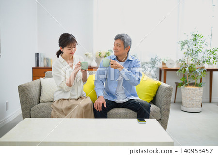 A smiling couple in their 60s sitting on a sofa and drinking coffee. 140953457