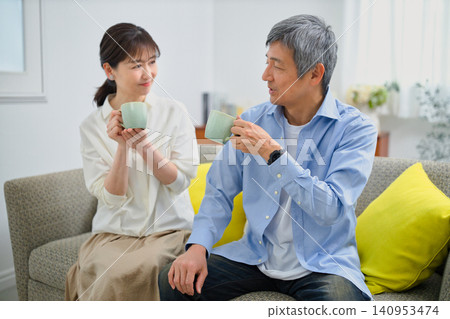 一對六十多歲的夫婦面帶笑容地坐在沙發上喝咖啡。 一對六十多歲的夫婦面帶笑容地坐在沙發上喝咖啡。 140953474