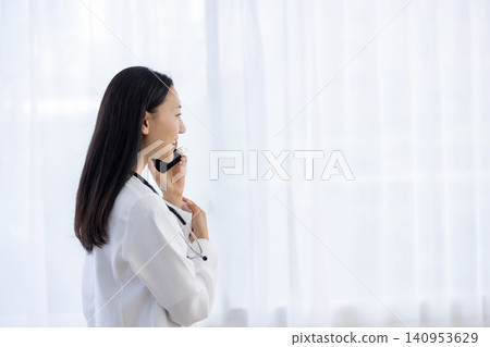 A Japanese female doctor smiles and talks while holding her smartphone to her ear by the window. A Japanese female doctor smiles and talks while holding her smartphone to her ear by the window. 140953629
