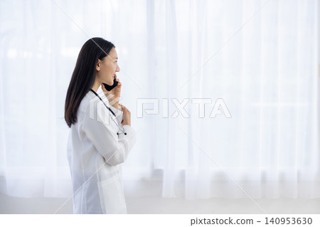 A Japanese female doctor smiles and talks while holding her smartphone to her ear by the window. A Japanese female doctor smiles and talks while holding her smartphone to her ear by the window. 140953630