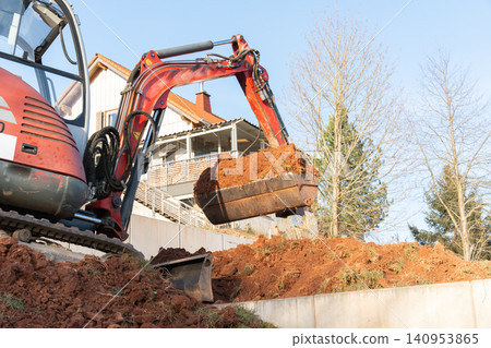 Mini-excavator in action during excavation works next to a residential house bucket filled with earth shot from below. High quality photo Mini-excavator in action during excavation works next to a residential house bucket filled with earth shot from below. High quality photo 140953865