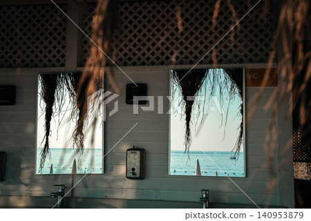 Beach scene reflected in two mirrors with hanging thatch framing the view. The serene ocean and beach umbrellas add to the relaxing coastal setting. Beach scene reflected in two mirrors with hanging thatch framing the view. The serene ocean and beach umbrellas add to the relaxing coastal setting. 140953879