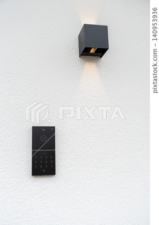 A stylish black smart doorbell with a keypad and camera mounted on a white textured wall below a modern black wall lamp emitting soft light. Ideal for home security and smart technology concepts A stylish black smart doorbell with a keypad and camera mounted on a white textured wall below a modern black wall lamp emitting soft light. Ideal for home security and smart technology concepts 140953936