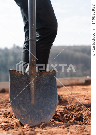 A black iron shovel is firmly stuck in the ground, standing upright at the feet of a worker. The image captures the essence of manual labor, outdoor work, and construction with a focus on tools and A black iron shovel is firmly stuck in the ground, standing upright at the feet of a worker. The image captures the essence of manual labor, outdoor work, and construction with a focus on tools and 140953938