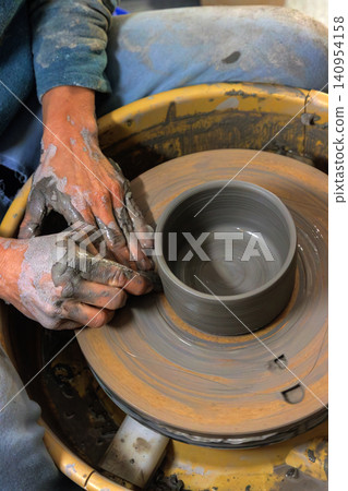 Clay covered hands shaping wet clay on a spinning pottery wheel in a ceramic studio. Selective focus. Motion effect. Clay covered hands shaping wet clay on a spinning pottery wheel in a ceramic studio. Selective focus. Motion effect. 140954158