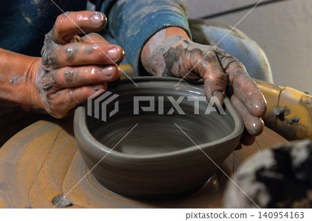 Clay covered hands shaping wet clay on a spinning pottery wheel in a ceramic studio. Selective focus. Motion effect. Clay covered hands shaping wet clay on a spinning pottery wheel in a ceramic studio. Selective focus. Motion effect. 140954163