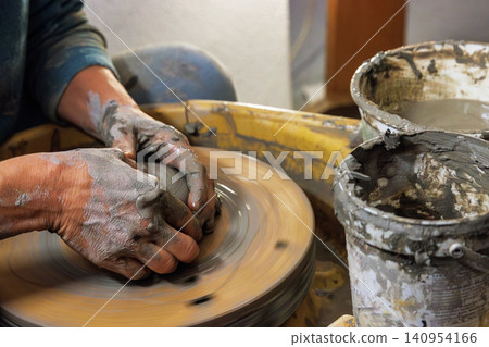 Closeup of clay covered hands shaping wet clay on a spinning pottery wheel in a ceramic studio. Selective focus. Motion effect. Closeup of clay covered hands shaping wet clay on a spinning pottery wheel in a ceramic studio. Selective focus. Motion effect. 140954166