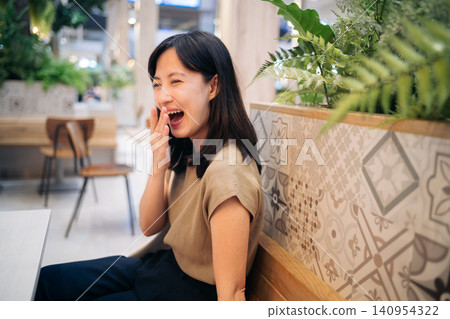 Smiling woman indoor casual clothing sitting wooden bench patterned tile green plant cheerful candid Asian relaxed modern cafe natural light Smiling woman indoor casual clothing sitting wooden bench patterned tile green plant cheerful candid Asian relaxed modern cafe natural light 140954322