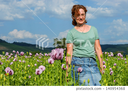 Smiling woman walking through a blooming pink poppy field in summer countryside, with green hills and cloudy blue sky in the background. 140954368