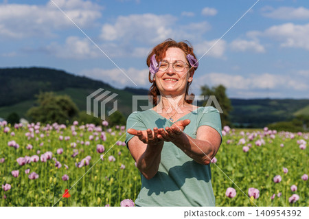Mature woman with glasses and curly red hair playfully throwing pink poppy petals while standing in a blooming poppy field. 140954392