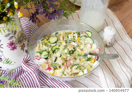 A bowl of okroshka filled with potatoes, chicken breast, cucumbers, herbs, and boiled eggs sits on a table. Nearby, a floral vase adds color to the setting A bowl of okroshka filled with potatoes, chicken breast, cucumbers, herbs, and boiled eggs sits on a table. Nearby, a floral vase adds color to the setting 140954977