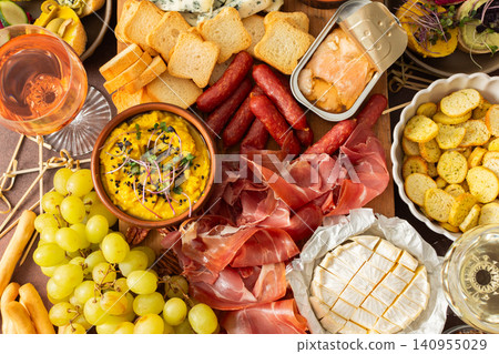 A vibrant charcuterie board showcases a variety of cheese, sausages, hummus, nuts, and sandwiches. Friends gather at a sunny outdoor picnic, enjoying delicious food together A vibrant charcuterie board showcases a variety of cheese, sausages, hummus, nuts, and sandwiches. Friends gather at a sunny outdoor picnic, enjoying delicious food together 140955029