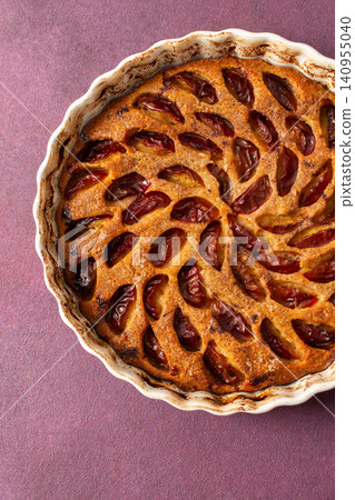 A plum pie is placed on a table with thinly sliced plums visible on top. Bowls of fresh plums are nearby with empty plates waiting for dessert A plum pie is placed on a table with thinly sliced plums visible on top. Bowls of fresh plums are nearby with empty plates waiting for dessert 140955040