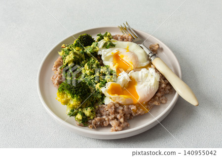 A healthy breakfast bowl features green buckwheat topped with pesto, steamed broccoli, and poached eggs. A fork rests beside the dish ready for a nutritious meal A healthy breakfast bowl features green buckwheat topped with pesto, steamed broccoli, and poached eggs. A fork rests beside the dish ready for a nutritious meal 140955042
