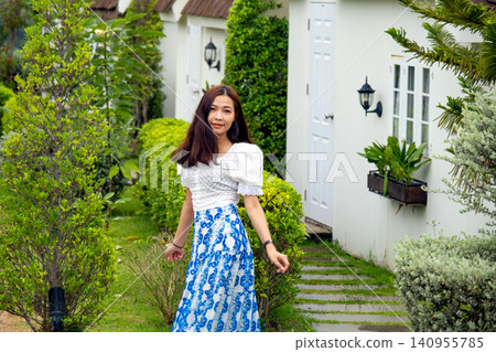 Smiling woman posing in a beautifully landscaped garden beside a charming white house, showcasing a peaceful outdoor lifestyle Smiling woman posing in a beautifully landscaped garden beside a charming white house, showcasing a peaceful outdoor lifestyle 140955785