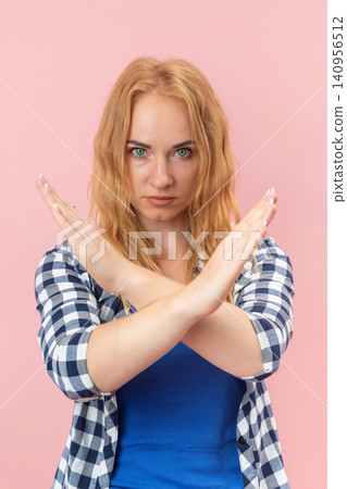 Young woman standing on pink background, crossing arms in front of her chest forming an X shape, expressing disagreement, rejection, or prohibition with a serious face Young woman standing on pink background, crossing arms in front of her chest forming an X shape, expressing disagreement, rejection, or prohibition with a serious face 140956512