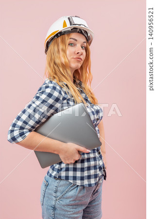 Skilled female professional builder or architect working in construction, wearing a safety helmet and plaid shirt, holding a modern laptop against a pink background Skilled female professional builder or architect working in construction, wearing a safety helmet and plaid shirt, holding a modern laptop against a pink background 140956521