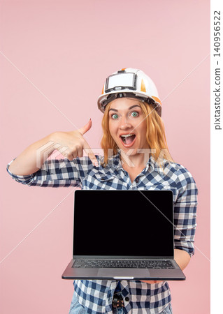 Excited woman worker wearing a hard hat and plaid shirt holding an open laptop, pointing down at the empty screen ready for custom advertising or business information Excited woman worker wearing a hard hat and plaid shirt holding an open laptop, pointing down at the empty screen ready for custom advertising or business information 140956522