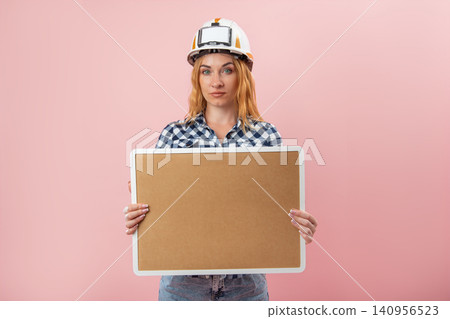 Confident woman construction worker is holding a blank cork board, presenting a clear message space for communication, advertising, or important announcements on a pink background Confident woman construction worker is holding a blank cork board, presenting a clear message space for communication, advertising, or important announcements on a pink background 140956523