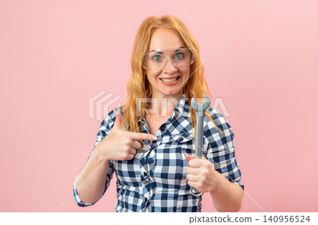 Happy young woman wearing plaid shirt and protective glasses holding a large wrench, pointing at herself with a confident and encouraging expression against a pink background Happy young woman wearing plaid shirt and protective glasses holding a large wrench, pointing at herself with a confident and encouraging expression against a pink background 140956524