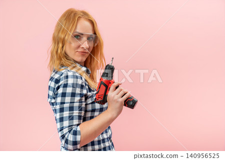 Young confident woman holding a red power drill, wearing a plaid shirt and safety glasses, representing female empowerment and do it yourself spirit Young confident woman holding a red power drill, wearing a plaid shirt and safety glasses, representing female empowerment and do it yourself spirit 140956525