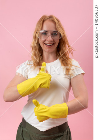 Smiling woman wearing protective safety glasses and yellow rubber gloves showing two thumbs up, indicating approval and excellent service on a pink background Smiling woman wearing protective safety glasses and yellow rubber gloves showing two thumbs up, indicating approval and excellent service on a pink background 140956537