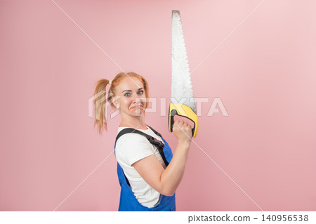 Young woman expressing confusion while holding a large hand saw, standing against a pink background, representing unexpected challenges or a task beyond her comfort zone Young woman expressing confusion while holding a large hand saw, standing against a pink background, representing unexpected challenges or a task beyond her comfort zone 140956538