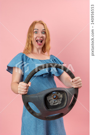 Young woman expressing surprise and excitement, holding a detached or broken car steering wheel missing its central cover, symbolizing mobility issues or unexpected road problems Young woman expressing surprise and excitement, holding a detached or broken car steering wheel missing its central cover, symbolizing mobility issues or unexpected road problems 140956553