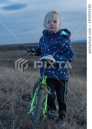 Little girl standing with her bright green bicycle in an open field, looking directly forward, capturing a moment of childhood exploration and nature connection 140956590